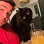 man, hat, red_shirt, black_cat, cat, table, glass, orange_juice, vape_device, indoor, wooden_beams, curious, companion, face, portrait, furniture, drinks, beverage, casual, cozy