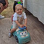 Loan a rejoint le concours — aidez-le/la à gagner de superbes lots ! toddler, child, ride_on_toy, blue, outdoor, smiling, play, toy_car, person, footwear, shorts, white_shirt, hands, tablecloth, flooring, adult, supervision, happy, blonde, eyes