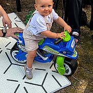 Loan a rejoint le concours — aidez-le/la à gagner de superbes lots ! toddler, child, toy_motorcycle, outdoor, grass, mat, person, footwear, hand, play, blue_eyes, shorts, white_shirt, plastic_toy, summer, daylight, cute, small_vehicle, fun, baby