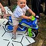 toddler, child, toy_motorcycle, outdoor, grass, mat, person, footwear, hand, play, blue_eyes, shorts, white_shirt, plastic_toy, summer, daylight, cute, small_vehicle, fun, baby