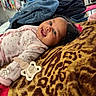 baby, blanket, casual, child, clothing, curly_hair, face, furniture, happy, home, indoors, lying_down, pacifier, pacifier_clip, patterned_blanket, person, pink, room, smiling, television