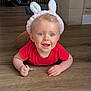 baby, blue_eyes, bunny_ears, child, crawling, cute, floor, hands, happy, headband, indoor, infant, light_blond_hair, person, playful, portrait, red_shirt, smiling, wooden_floor, young_child