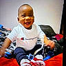 baby, child, smiling, tshirt, clothing, sitting, blanket, red, blue, black, white, face, happy, indoor, person, footwear, pants, toy, bed, casual
