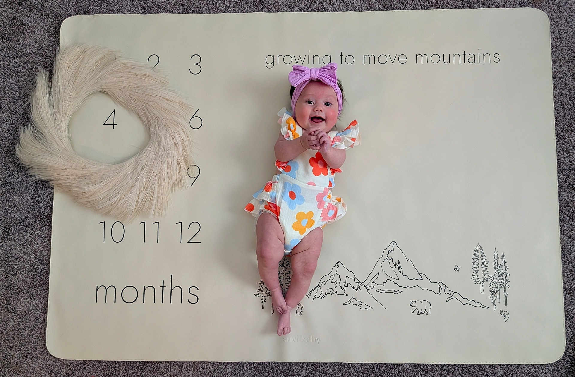 Sosie is registered to the contest to win money with this photo: baby, infant, smiling, headband, purple_bow, floral_outfit, milestone_mat, months, numbers, mountains, carpet, floor, happy, cute, child, indoor, lying_down, celebration, growth, portrait