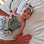 baby, infant, child, hand, bed, white_bed, bow, headband, green_top, floral_skirt, person, skin, fingers, lying_down, indoors, soft_light, adult_hand, curious, cute, portrait