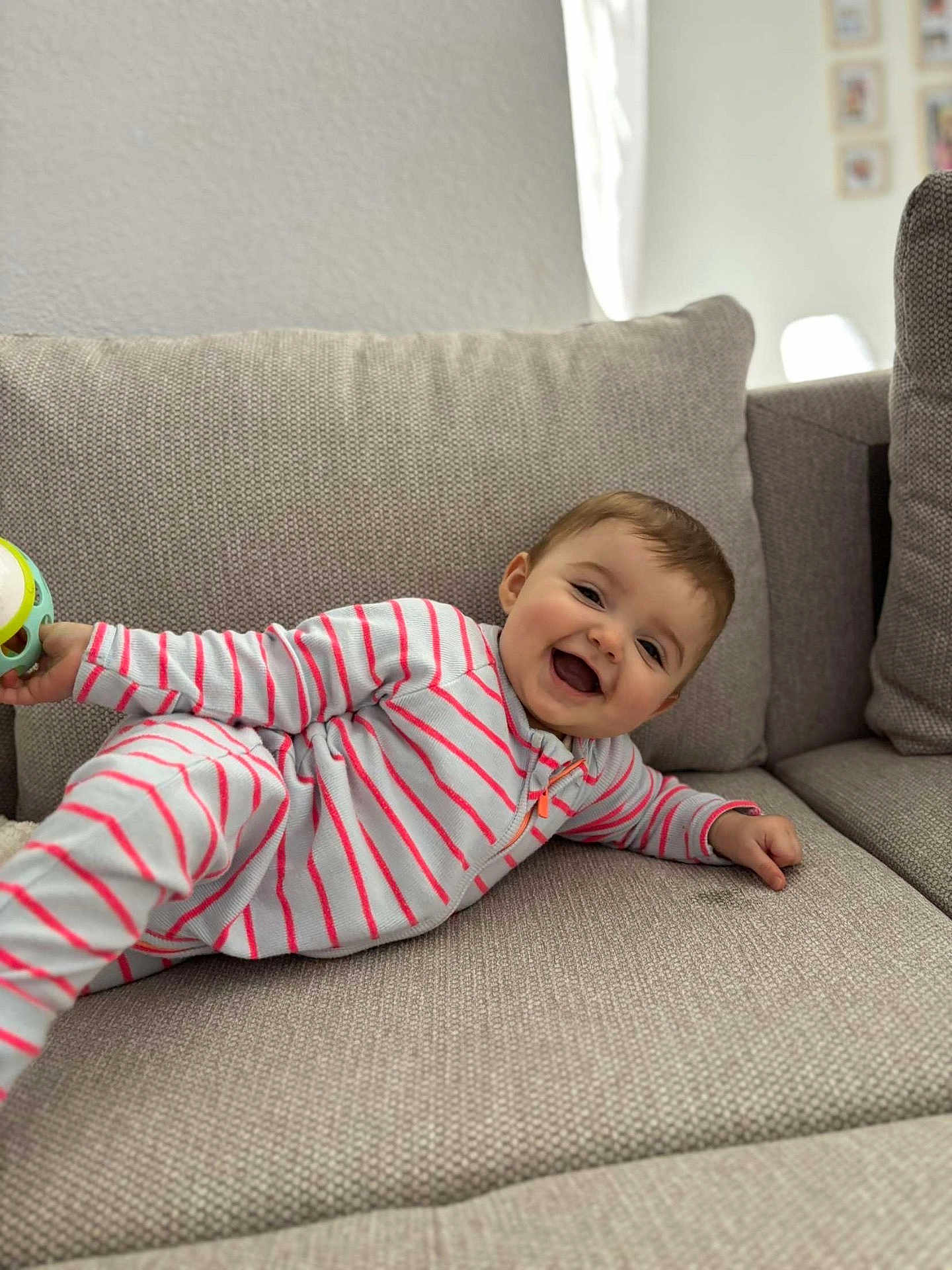 Alba participe au concours pour gagner de l'argent avec cette photo : baby, child, smiling, striped_onesie, couch, living_room, toy, happy, infant, face, person, indoor, cozy, furniture, sitting, cute, playful, comfort, portrait, cushion