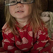 Ashley is registered to the contest to win money with this photo: child, glasses, pajamas, heart_pattern, red_clothing, sitting, indoors, couch, blanket, cozy, curly_hair, face, person, comfort, playful, smiling, young, cute, home, soft