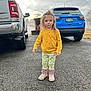 Ashley is registered to the contest to win money with this photo: child, toddler, yellow_sweater, patterned_pants, leopard_boots, standing, driveway, asphalt, jeep, pickup_truck, vehicles, overcast_sky, clouds, parking, rosy_cheeks, portrait, outdoors, reflection, garage, trees