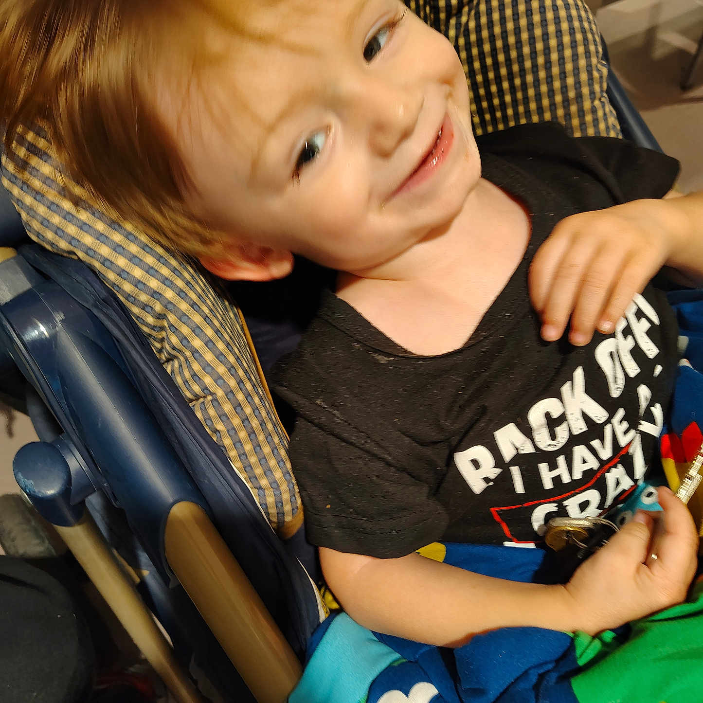 Charles is registered to the contest to win money with this photo: black_shirt, blanket, blue, casual, child, clothing, colorful, cute, face, hand, happy, indoor, pattern, person, seat, smiling, stroller, text_on_shirt, toddler, young