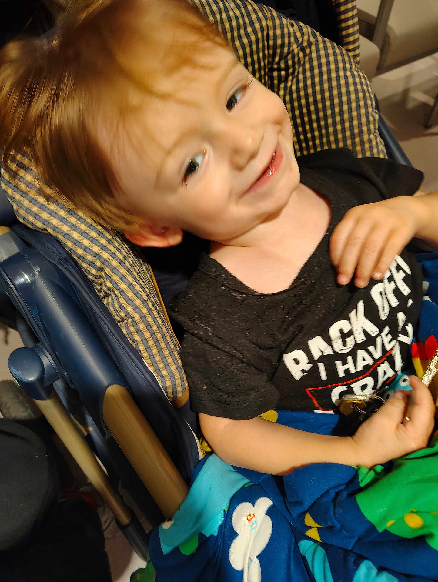 Charles is registered to the contest to win money with this photo: child, smiling, toddler, stroller, blanket, clothing, indoor, face, hand, person, text_on_shirt, colorful, blue, black_shirt, happy, cute, young, seat, pattern, casual