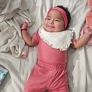 Azriella is registered to the contest to win money with this photo: baby, infant, smile, pink_clothing, headband, bed, blanket, lying_down, cute, child, person, indoors, soft_fabric, happy, portrait, babies, toddler, cozy, sleepwear, resting