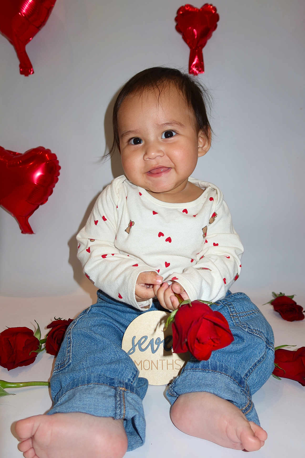 Damian joined the competition — help win amazing prizes! baby, child, rose, red_rose, heart_balloon, balloon, jeans, white_background, barefoot, smiling, portrait, milestone, seven_months, long_sleeve_shirt, heart_pattern, cute, sitting, person, indoor, celebration