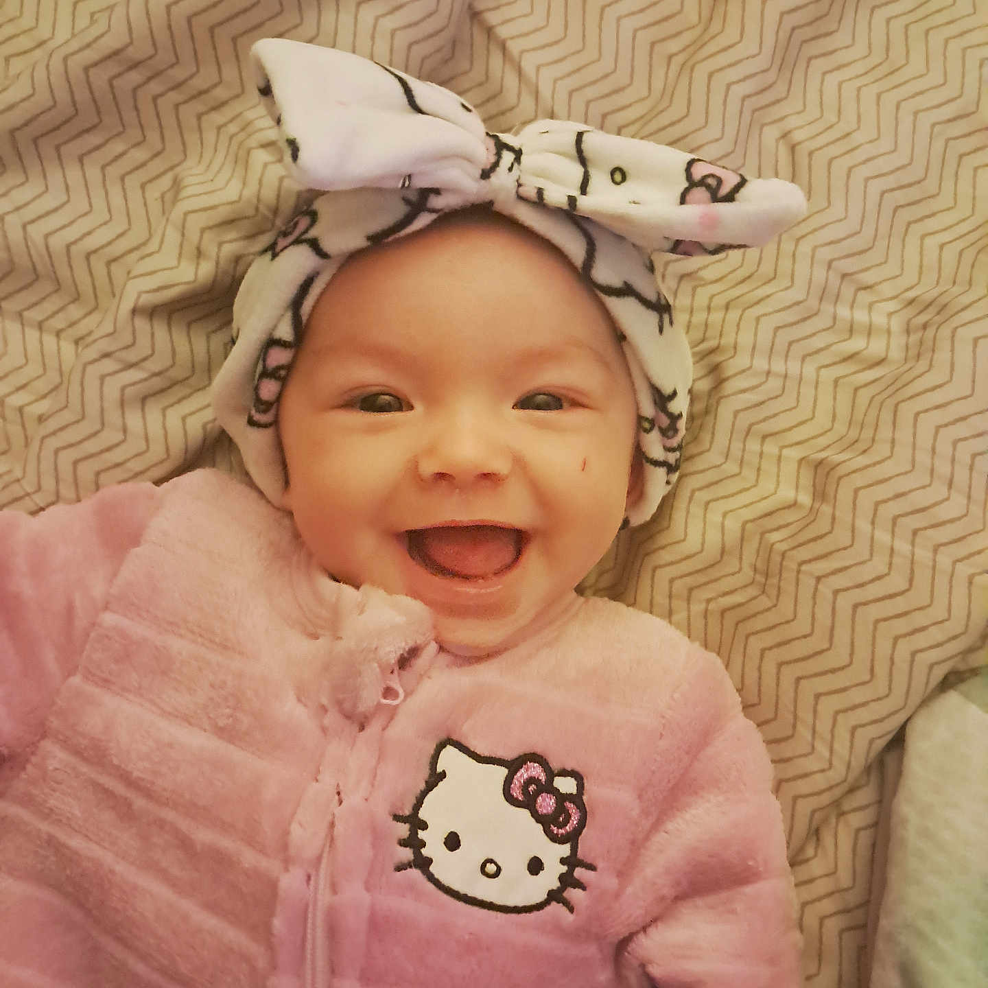 Raya is registered to the contest to win money with this photo: apparel, baby, blanket, bow, child, cozy, cute, face, happy, headband, hello_kitty, indoors, infant, lying_down, pattern, pink_clothing, portrait, smile, warm_clothing, young_child