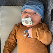 Julius is registered to the contest to win money with this photo: baby, infant, child, portrait, pacifier, blue_hat, brown_shirt, clothing, sitting, cushion, seat, indoor, close_up, face, eyes, hand, furniture, cozy, calm, photography