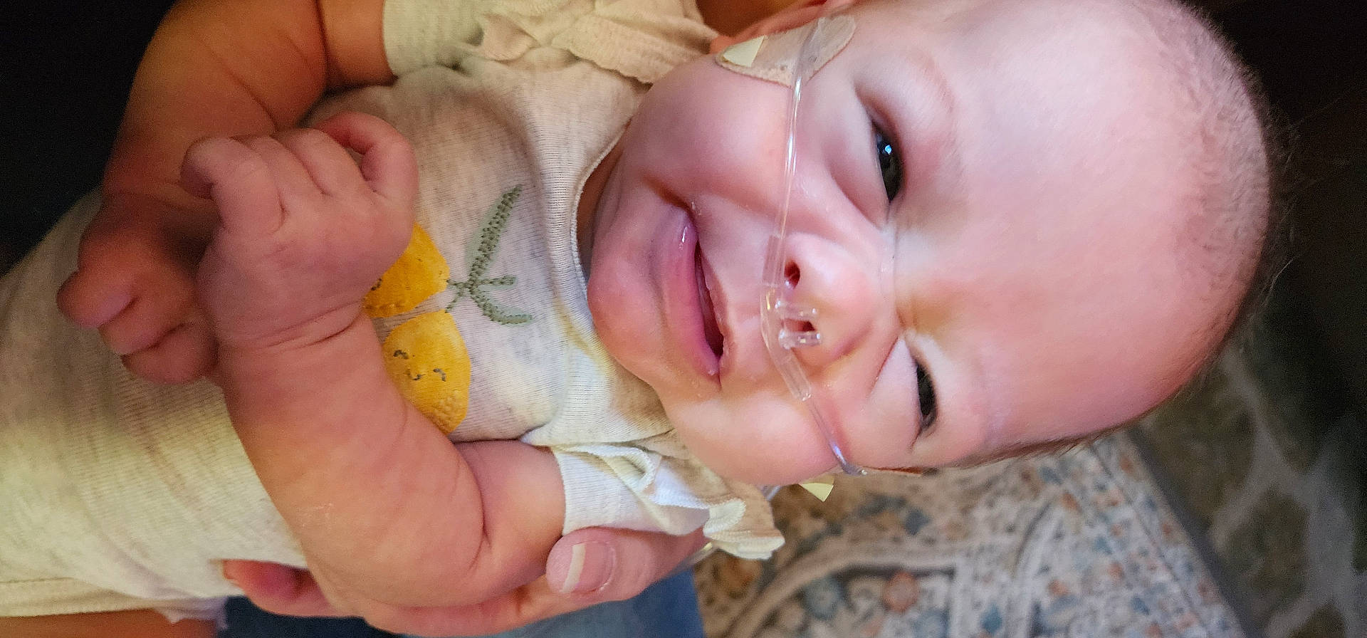 arm, baby, carpet, closeup, clothing, cute, expression, face, hand, healthcare, holding, indoor, infant, medical, nasal_cannula, newborn, pattern, person, skin, smile