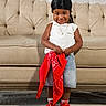 toddler, child, smiling, standing, red_sneakers, black_durag, white_top, denim_skirt, red_bandana, beige_couch, indoor, portrait, happy, cute, person, floor_covering, jewelry, necklace, fashion, casual