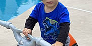 Cameron is registered to the contest to win money with this photo: toddler, child, sunglasses, toy_motorcycle, pool, backyard, blue_shirt, dinosaur_print, gray_pants, outdoor, concrete, footwear, playing, summer, sunny, young_child, vehicle, riding, casual_clothing, fun