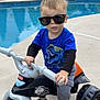 toddler, child, sunglasses, toy_motorcycle, pool, backyard, blue_shirt, dinosaur_print, gray_pants, outdoor, concrete, footwear, playing, summer, sunny, young_child, vehicle, riding, casual_clothing, fun
