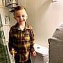 child, boy, smiling, plaid_shirt, toy, bathroom, toilet, shower_curtain, shelf, bottle, skin_care, soap, person, indoor, casual_clothing, short_hair, happy, standing, home, portrait