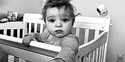 Coralie is registered to the contest to win money with this photo: baby, crib, child, indoor, monochrome, person, hand, face, curly_hair, expression, bedroom, door, blanket, toy, wall, carpet, long_sleeve, furniture, infant, looking