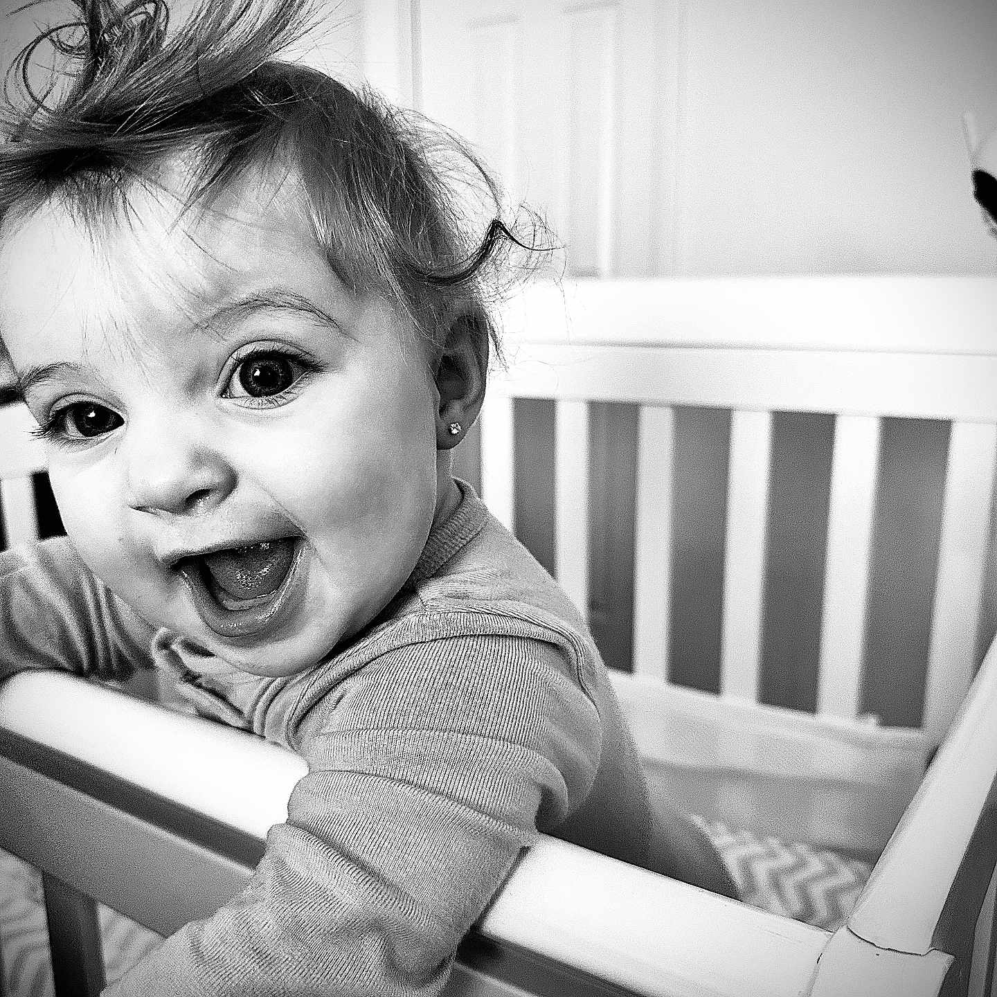 Coralie is registered to the contest to win money with this photo: baby, black_and_white, child, clothing, crib, cute, earring, expression, face, hair, happy, home, indoor, infant, person, playful, portrait, railing, smiling, young_child