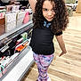 child, girl, curly_hair, black_shirt, butterfly, colorful_pants, galaxy_print, white_shoes, store, stationery, notebooks, wooden_floor, pose, smile, playful, aisle, shelf, shopping, indoor, person