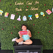 Pheonix is registered to the contest to win money with this photo: child, smiling, sitting, greenery, backdrop, school, sign, love, shoes, shorts, tank_top, platform, decorations, happy, cute, cute_child, playful, outdoor, fun, celebration