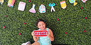 Pheonix is registered to the contest to win money with this photo: backdrop, celebration, child, cute, cute_child, decorations, fun, greenery, happy, love, outdoor, platform, playful, school, shoes, shorts, sign, sitting, smiling, tank_top