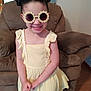 child, girl, sunglasses, yellow_dress, smiling, hairstyle, buns, indoor, armchair, brown_furniture, happy, portrait, person, fashion, casual, cute, young, wooden_floor, home, fun