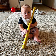 Cade is registered to the contest to win money with this photo: baby, child, carpet, indoor, toy, bat, yellow, plastic, fan, balloon, stuffed_toy, floor, person, sitting, curious, black_shirt, shorts, bib, home, play