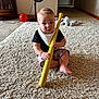 baby, child, carpet, indoor, toy, bat, yellow, plastic, fan, balloon, stuffed_toy, floor, person, sitting, curious, black_shirt, shorts, bib, home, play