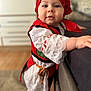 toddler, child, red_headscarf, traditional_clothing, white_lace, standing, indoor, carpet, couch, curious_expression, face, person, cute, young_child, colorful_outfit, home_interior, soft_focus, floor, holding, portrait