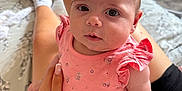 Blue is registered to the contest to win money with this photo: baby, child, infant, pink_dress, floral_pattern, hand, person, indoor, bed, blanket, socks, furniture, drawer, carpet, skin, cute, portrait, sitting, looking_at_camera, human