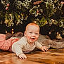 baby, child, smiling, lying_down, floor, wooden_floor, christmas_tree, holiday, festive, decorations, lights, pine_branches, red_hair, striped_pants, cream_sweater, indoors, happy, cute, seasonal, celebration