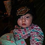baby, child, face, hat, beanie, clothing, blanket, patterned, harley_davidson, indoor, person, drink_cup, dark_background, curious_expression, seated, warm_clothing, cute, portrait, infant, casual