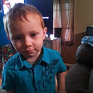 Aaron is registered to the contest to win money with this photo: child, boy, blue_shirt, indoor, living_room, television, curtains, furniture, person, portrait, young_child, casual_clothing, home_interior, curious, standing, hand_on_table, soft_lighting, red_hair, face, expression