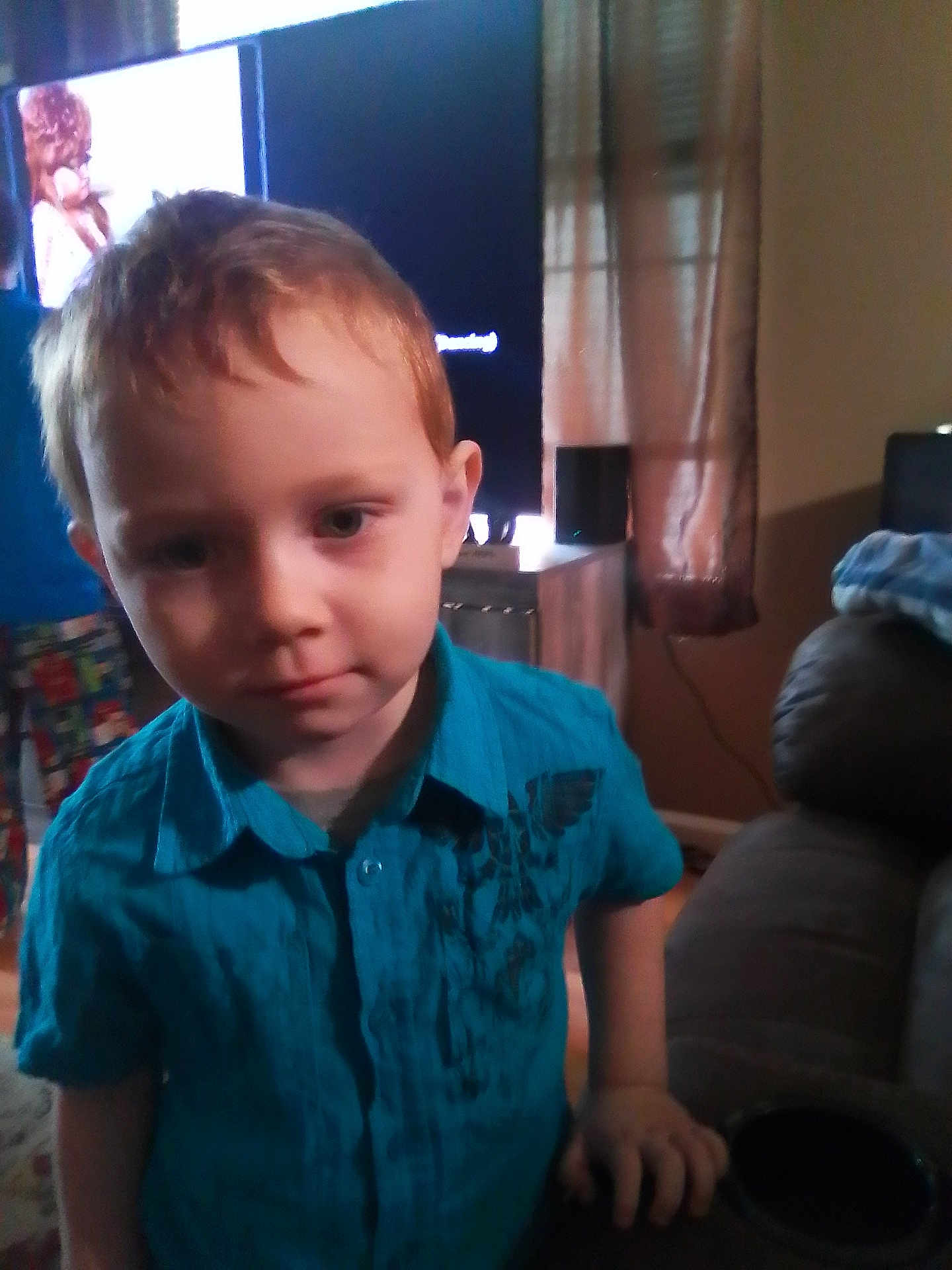 Aaron is registered to the contest to win money with this photo: child, boy, blue_shirt, indoor, living_room, television, curtains, furniture, person, portrait, young_child, casual_clothing, home_interior, curious, standing, hand_on_table, soft_lighting, red_hair, face, expression