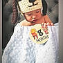 Oscar is registered to the contest to win money with this photo: baby, infant, crochet_hat, puppy_hat, blanket, milestone_sign, drooling, couch, tufted_furniture, wrapped, cute, child, indoors, soft_texture, white_blanket, seated, portrait, headwear, adorable, young_child