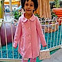 Connie joined the competition — help win amazing prizes! child, pink_coat, handbag, curly_hair, white_fence, indoor, shopping_mall, play_area, plants, decorative_tree, sneakers, standing, portrait, young_person, curious_expression, background_people, flooring, green_decor, buttoned_coat, casual_clothing
