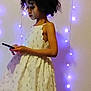 child, dress, curly_hair, smartphone, fairy_lights, purple_lights, wall, side_profile, standing, indoor, portrait, candid, bedroom, soft_lighting, app_ui, screenshot, patterned_dress, holding_phone, young_person, decor
