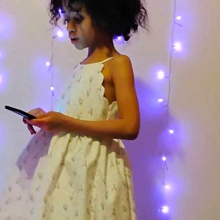 Marni is registered to the contest to win money with this photo: app_ui, bedroom, candid, child, curly_hair, decor, dress, fairy_lights, holding_phone, indoor, patterned_dress, portrait, purple_lights, screenshot, side_profile, smartphone, soft_lighting, standing, wall, young_person