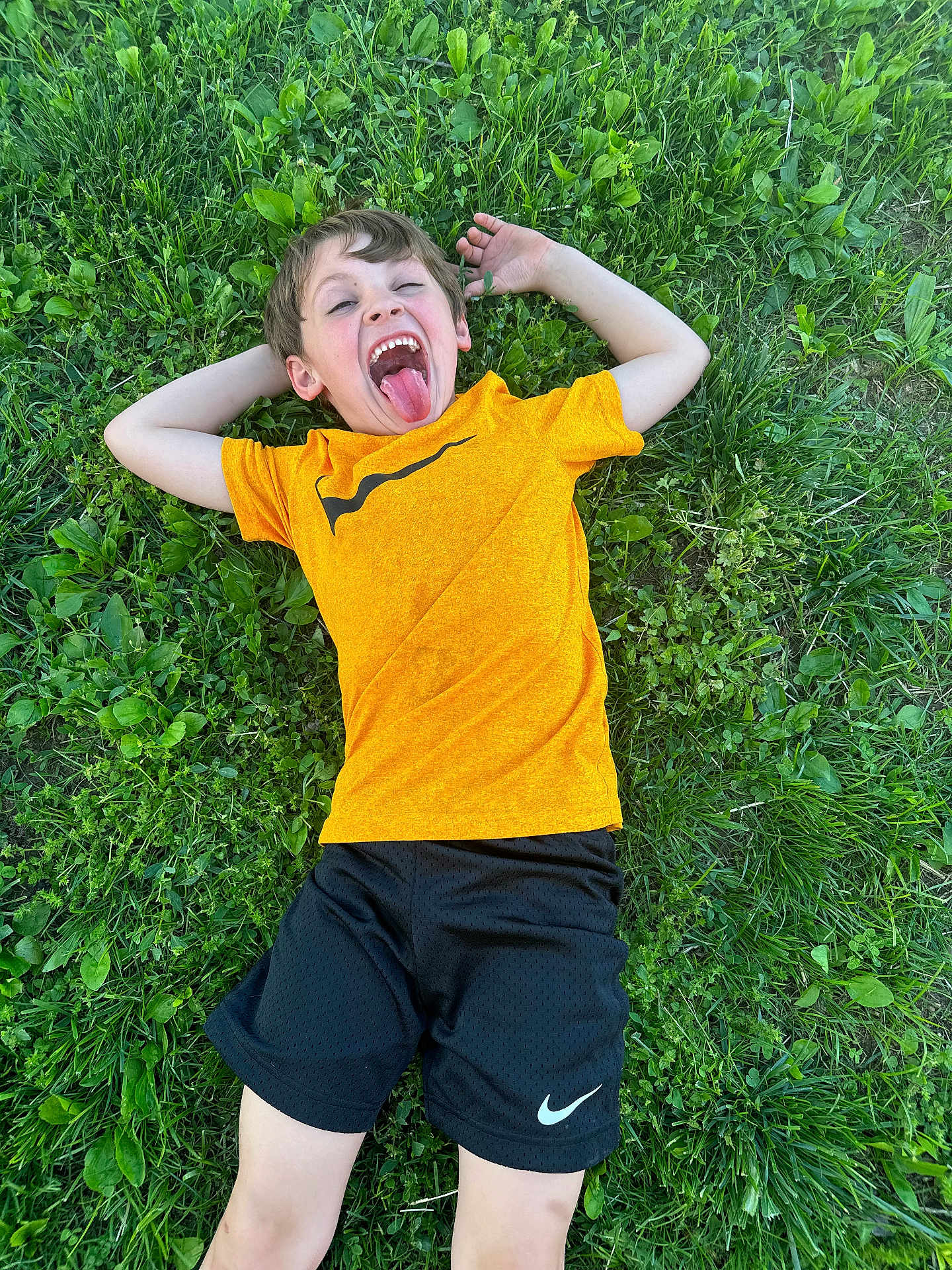 Brycen is registered to the contest to win money with this photo: child, boy, grass, greenery, outdoor, playful, tongue_out, casual_clothing, orange_shirt, black_shorts, nature, smiling, happy, relaxed, summer, youth, person, fun, carefree, leisure