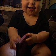 Sage is registered to the contest to win money with this photo: baby, child, infant, black_onesie, indoor, sitting, purple_object, television, people_on_tv, dim_lighting, smiling, cute, chubby_cheeks, person, furniture, home, background, relaxed, holding, candid