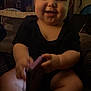 baby, child, infant, black_onesie, indoor, sitting, purple_object, television, people_on_tv, dim_lighting, smiling, cute, chubby_cheeks, person, furniture, home, background, relaxed, holding, candid