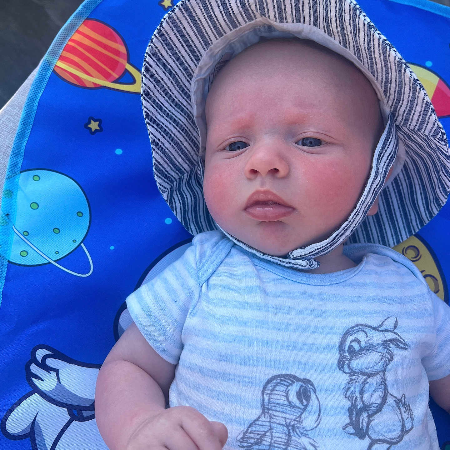 Enzo is registered to the contest to win money with this photo: baby, blue_shirt, cartoon_characters, child, clothing, colorful_mat, cute, disney_classics, expression, face, hand, hat, infant, outdoor, person, portrait, space_theme, striped_hat, sunhat, young