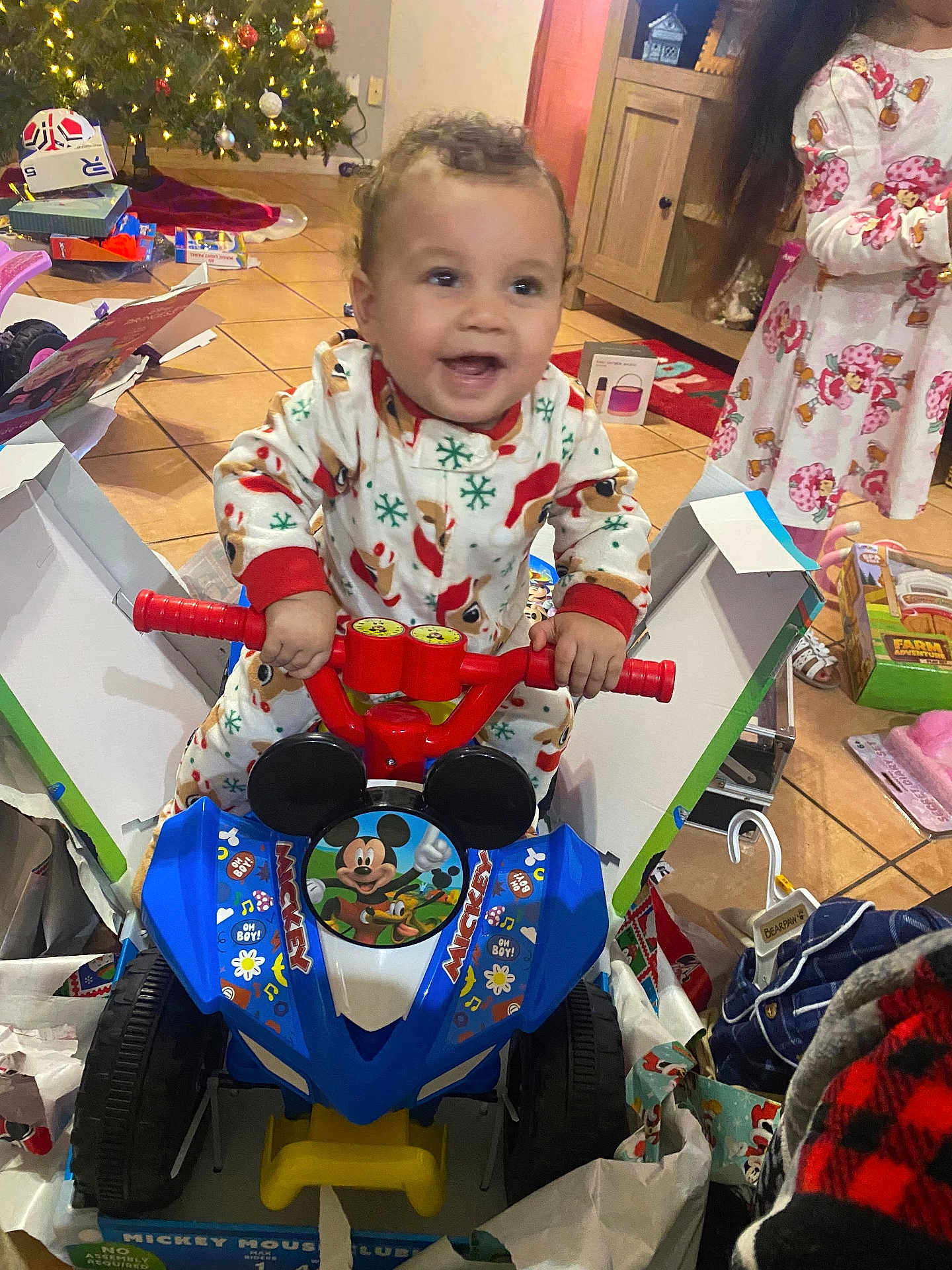 Jaden is registered to the contest to win money with this photo: toddler, child, toy_vehicle, mickey_mouse, pajamas, indoor, christmas_tree, holiday, wrapping_paper, gift_box, floor_tiles, cabinet, smiling, playtime, festive, toy, person, joyful, unwrapping, family