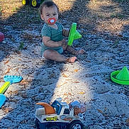 Jaden is registered to the contest to win money with this photo: toddler, child, sand, toy, toy_truck, dinosaur_toy, green_toy, pacifier, outdoor, play_area, grass, leaves, sunlight, shadow, plastic_toy, barefoot, short_sleeve, curly_hair, nature, playtime