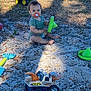 toddler, child, sand, toy, toy_truck, dinosaur_toy, green_toy, pacifier, outdoor, play_area, grass, leaves, sunlight, shadow, plastic_toy, barefoot, short_sleeve, curly_hair, nature, playtime