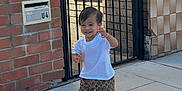 Sahas is registered to the contest to win money with this photo: toddler, child, smiling, white_tshirt, patterned_pants, sandals, sidewalk, brick_wall, iron_gate, outdoor, happy, person, walking, sunlight, playful, young_child, casual_clothing, urban, daytime, cute