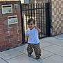 toddler, child, smiling, white_tshirt, patterned_pants, sandals, sidewalk, brick_wall, iron_gate, outdoor, happy, person, walking, sunlight, playful, young_child, casual_clothing, urban, daytime, cute